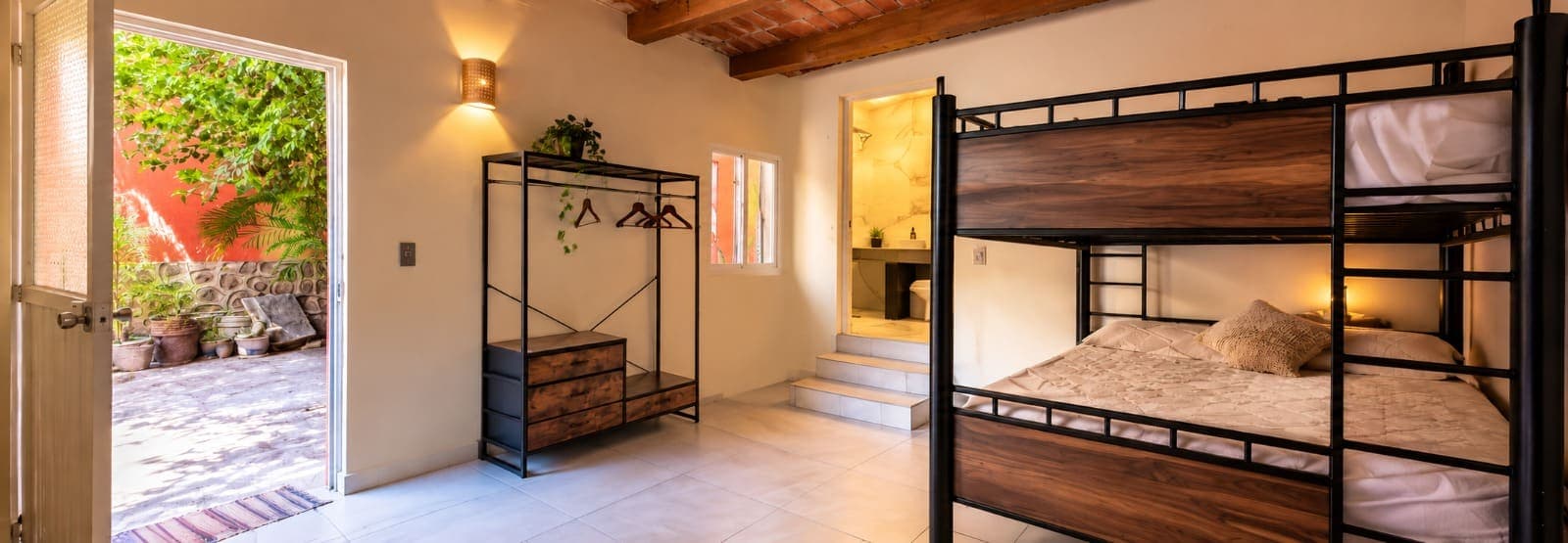 Un cuarto de la casa con litera de madera y herrería, perchero abierto, el baño al fondo y la puerta abierta al patio con plantas