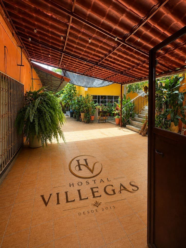 La entrada de la casa: un corredor con techo de teja, piso de loseta con el logotipo del hostal, helechos y macetas al fondo y la escalera que sube a la terraza