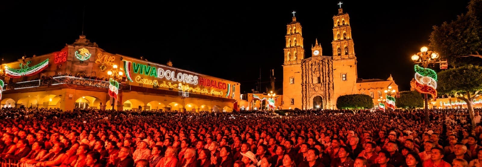 El Jardín Principal de noche en fiestas patrias: la parroquia iluminada, la presidencia con el letrero «Viva Dolores Hidalgo» y la plaza llena de gente