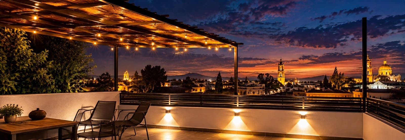 La terraza de la casa al anochecer: la pérgola con las luces encendidas, sillas junto al barandal y las torres del centro iluminadas al fondo