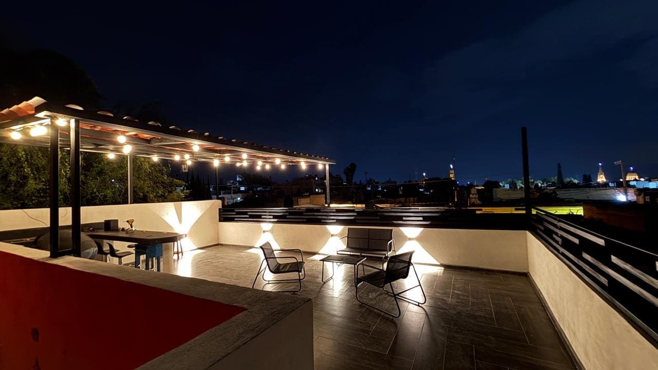 La terraza de noche: las luces colgadas de la pérgola encendidas, sillones bajos alrededor de una mesa y el pueblo iluminado al fondo